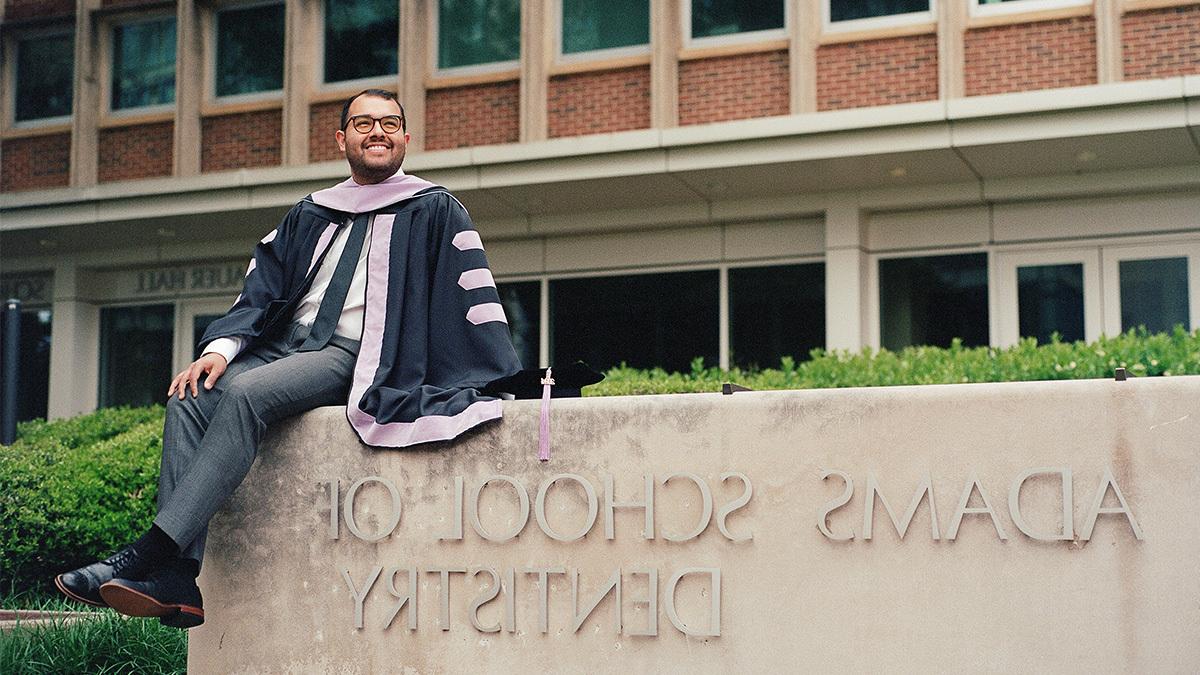 Bruno Segovia-Chumbez wearing commencement robes and sitting on wall reading 